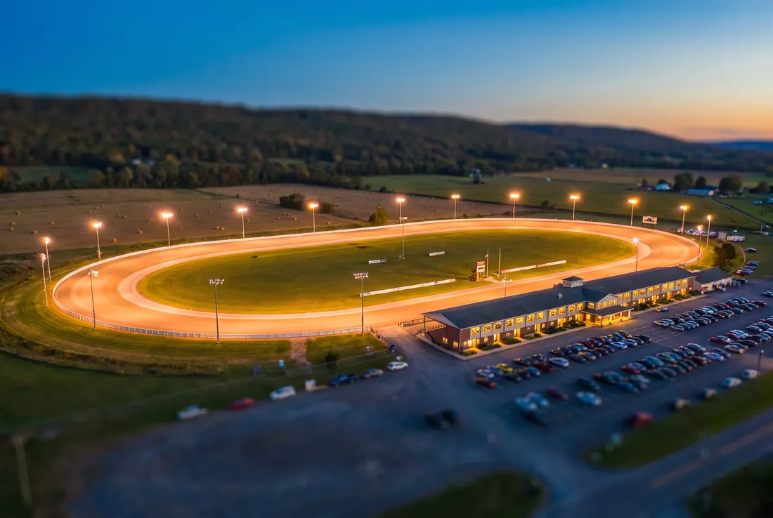 Vue miniature façon tilt-shift d'un ovale de courses attelées éclairé au crépuscule, avec une longue tribune et des terres agricoles au loin