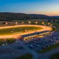 Vue miniature façon tilt-shift d'un ovale de courses attelées éclairé au crépuscule, avec une longue tribune et des terres agricoles au loin